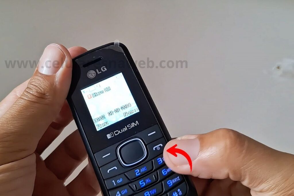 Mãos segurando um celular LG B220 preto. Um polegar pressiona o botão de encerrar chamada/liga/desliga, marcado por uma seta vermelha. A tela está acesa, mostrando a rede e data.